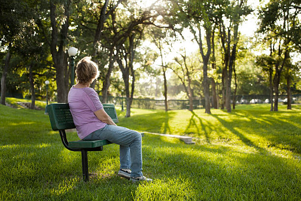 Rear view of mature woman sitting on park bench.  Summer. Rear view of one mature woman sitting on a park bench in the summer season.  Bright sunny day with lush green grass and trees.  The woman gazes off into the distance as she relaxes on a beautiful day.  Solitude, lonliness, contemplation.  She has short blond hair and wears a purple shirt and jeans.   Copyspace to right in this tranquil nature scene. bench stock pictures, royalty-free photos & images