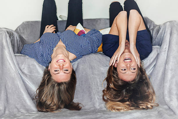 Two smiley girls laying upside down on a grey sofa stock photo