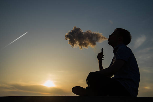 vaping young man with, produces vapor on sunset sky background at the sea coast promenade, place for text.