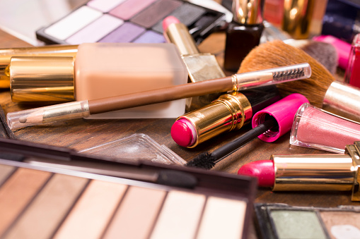 Large pile of various cosmetics lie on top of a wooden dressing table, vanity, or desk.  Make-up items include: eye shadow, nail polish, foundation, lipstick, make-up brushes,  blush, mascara, lip gloss.  Make-up artists' tools.  Great background.  Copyspace in center, top.