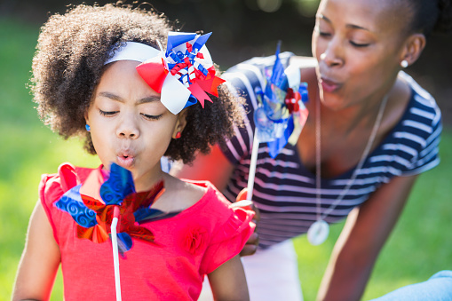An African American mother with her mixed race daughter celebrating an American patriotic holiday, perhaps July 4th or Memorial Day. They are playing with red, white and blue pinwheels, blowing on them. The focus is on the little girl, who is part black, Asian and Hispanic.