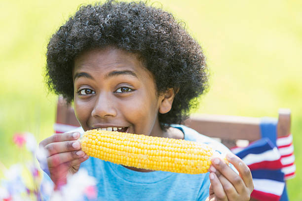 Face of a 12 year old mixed race boy, part black, Asian and Hispanic, enjoying an ear of corn at a July 4th or Memorial Day picnic. He is smiling at the camera.