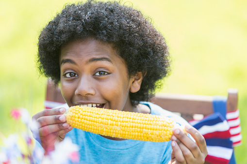 Face of a 12 year old mixed race boy, part black, Asian and Hispanic, enjoying an ear of corn at a July 4th or Memorial Day picnic. He is smiling at the camera.