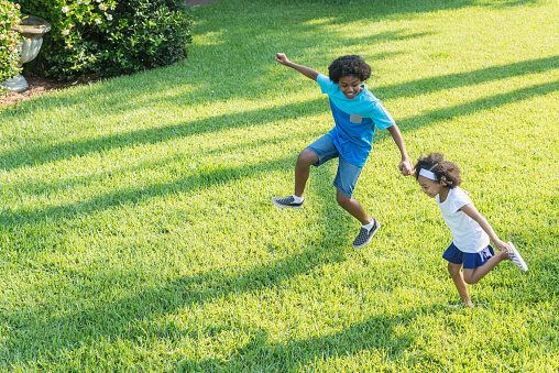 A 12 year old boy playing with his 7 year old sister in the park on a sunny day, holding her hand as they run and skip across the grass, smiling. They are mixed race, part African American, Hispanic, Asian and Pacific Islander ethnicity.