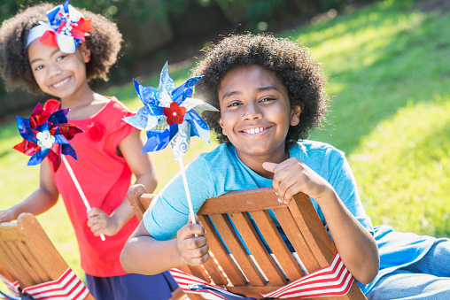 Two mixed race children having fun playing at an American patriotic event, with red, white and blue pinwheels. It could be a Memorial Day or 4th of July holiday cookout. The focus is on the 12 year old boy smiling at the camera. His 7 year old sister is out of focus in the background. They are part black, Hispanic and Asian.
