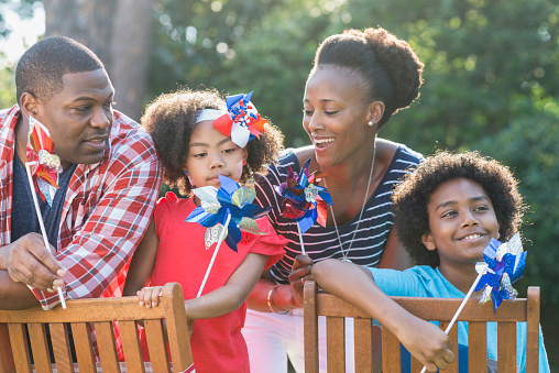 A mixed race African American family of four outdoors celebrating an American patriotic holiday, perhaps Memorial Day or July 4th. They are playing with red, white and blue pinwheels. The children are part black, Asian and Hispanic.