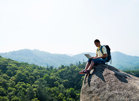 Asian male hiker using laptop on the top of mountain.