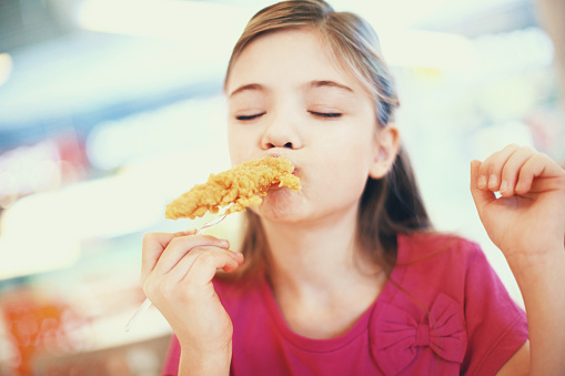 Closeup front view of cute little girl eating fried chicken and yes,enjoying it. She has a piece of chicken stuck on a fork and biting it with pleasure. Her eyes are closed, the girl is wearing pink t-shirt.