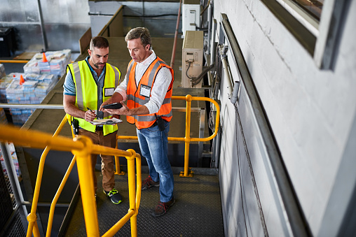 Shot of two warehouse workers standing on stairs discussing papework