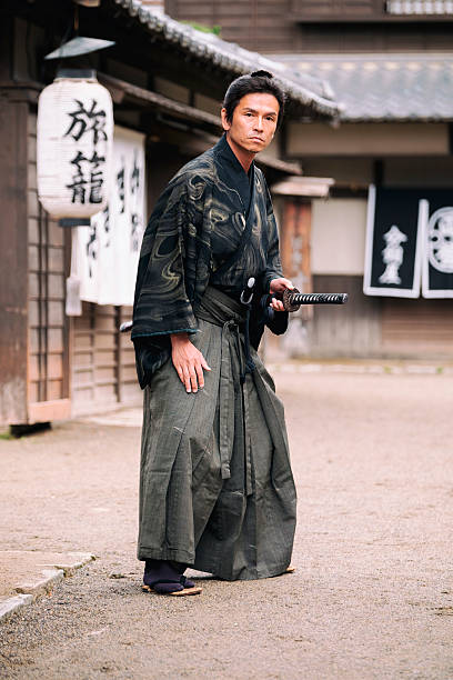 A Japanese Ronin Samurai holds his sword and faces the camera in a defensive posture. Standing on a dirt street in an Edo Period town. Photographed on location in Kyoto, Japan.