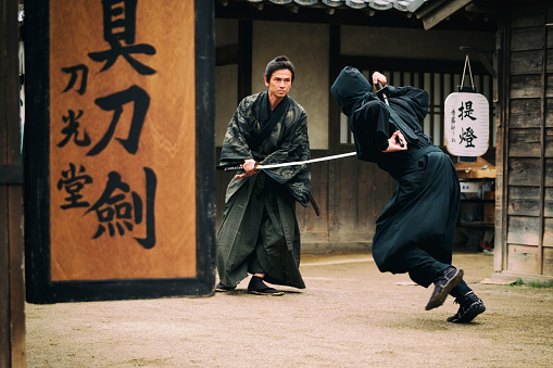 A Japanese Ronin Samurai holds his sword and battles a Ninja. Standing on a dirt street in an Edo Period town. Photographed on location in Kyoto, Japan.