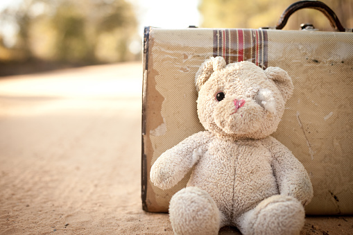 Color stock photo of an old, ragged teddy bear by a suitcase at the side of a dirt road in the rural country.