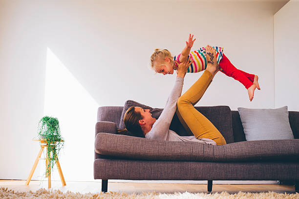 She is Flying High! Mother balancing her little girl on her feet as she lays on the sofa on her back. The little girl is pretending to fly. Both are happy superhero photos stock pictures, royalty-free photos & images