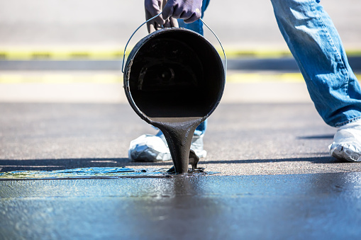 Close-up of a man pouring black tar seal coating onto the driveway as part of a do it yourself home improvement project. Taken on a summer day. Yellow 