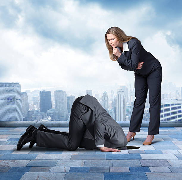 Businesswoman Looking At Businessman Who Has Head Buried In Hole A businesswoman looks down with concern at a businessman who is crouching on all fours as he buries his head in a hole.  She is concerned that he is avoiding the responsibilities of his job.  In the background is the skyline of a major urban city. head in the sand stock pictures, royalty-free photos & images