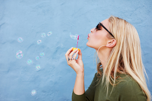Side view shot of attractive female model blowing soap bubbles. Young woman having fun against blue background.