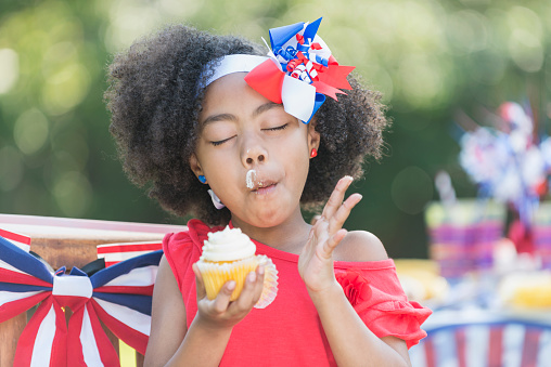A cute little mixed race 7 year old girl enjoying a cupcake at a July 4th or Memorial Day picnic. She is part Hispanic, Asian and black. The table and chair behind her are decorated in red, white and blue, celebrating American patriotism.