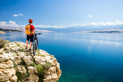 Mountain biking rider with bike looking at inspiring sea and mountains landscape. Man cycling MTB on enduro rocky trail path at sea side. Summer sport, training fitness motivation and inspiration.