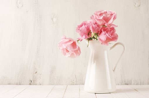 Fresh tender pink garden roses in white jug on rustic white wooden background. Filtered retro stylized image.