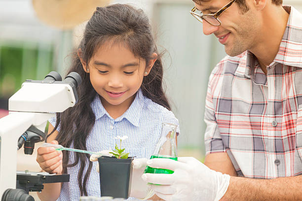 Happy girl watering a plant with her science teacher Young hispanic girl with her caucasian male teacher. She has long brown hair and is  using a pipette to measure out green liquid from the beaker. He is wearing glasses and plastic gloves. There is a microscope and small plants in front of them. biology class stock pictures, royalty-free photos & images