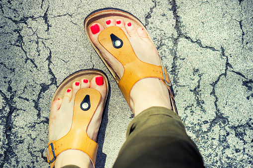 Woman's feet in yellow stylish summer sandals with red nail polish, standing on a cracked pavement on a warm summer day.