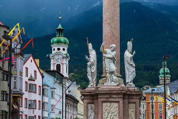 A view along Maria-Theresien-Strasse in Innsbruck during the day. The Annasaule, Hospital Church and other buildings can be seen. Mountains can be seen in the distance.