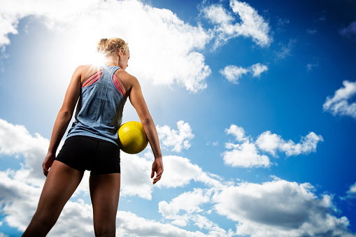 This is a photo of the back of a teenage female athlete holding a ball on a beach volleyball court in a cloudscape setting. This photo is taken from a low angle capturing a great clouds in front of her and sun flare coming off her shoulder. She is looking out into the horizon.