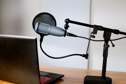 Horizontal image of audio equipment and a laptop sitting on a desk in a small sound room. The studio microphone is on a boom and has a pop filter in place. The laptop is open and ready to use.