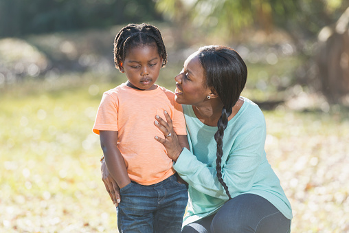 A young African American mother trying to cheer up her sad little boy, who is looking down with a sad face, hands in his pockets. She is kneeling beside him, arm around to comfort him, smiling and talking.