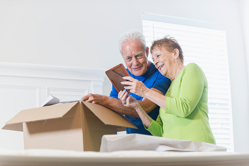 A senior couple moving house, packing a cardboard box in the bedroom. They are talking and smiling, looking at a picture frame which is bringing back memories.