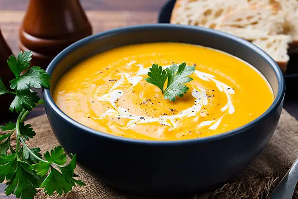 Pumpkin and carrot soup with cream and parsley on dark wooden background.