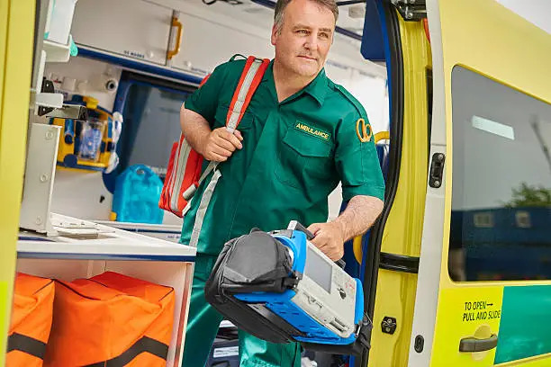 a uk ambulance staff member emerges from the back of an ambulance with his emergency backpack , and vital signs monitor .