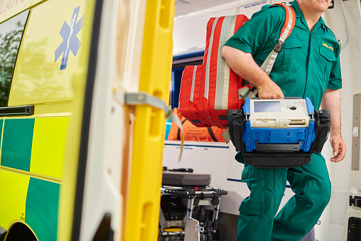 a uk ambulance staff member emerges from the back of an ambulance with his emergency backpack , and vital signs monitor .