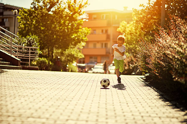 Little boy playing soccer on the street Little boy is playing football in the city. The boy aged 6 is running after the football and smiling. Sunny summer day in city residential area. center athlete stock pictures, royalty-free photos & images