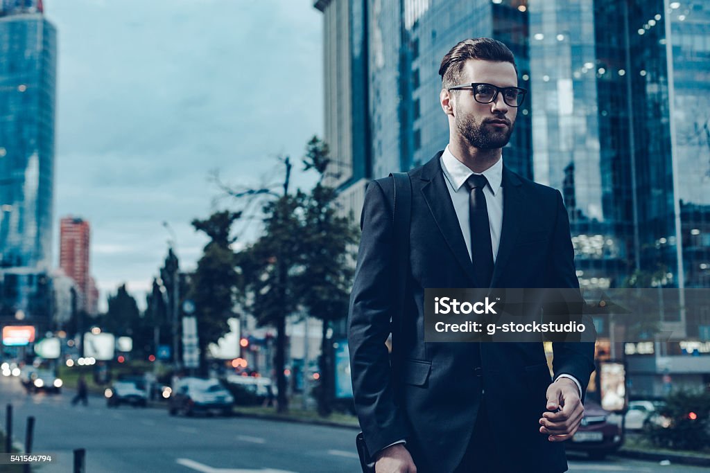 Businessman on the go. Night time image of confident young and handsome man in full suit walking along the street with cityscape in the background 20-29 Years Stock Photo
