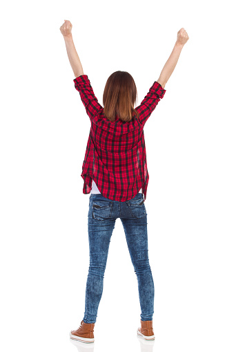Young woman in red lumberjack shirt, jeans and brown sneakers standing with arms raised. Rear view. Full length studio shot isolated on white.