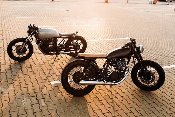 Two black and silver vintage custom motorcycles cafe racers Two cool vintage custom motorbike cafe racer looking in opposite directions on empty rooftop parking lot with backlight sun during sunset. Urban style, hipster lifestyle. cafe racer stock pictures, royalty-free photos & images