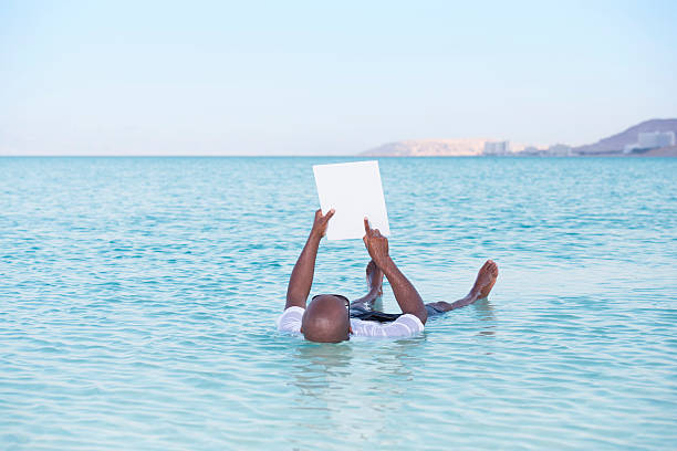 Tourist on vacation in Dead Sea. Unrecognisable bald man floating on water, touching on white paper blank.