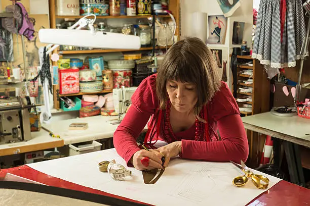 A woman tailor in red blouse and necklaces tracing and measuring white paper.