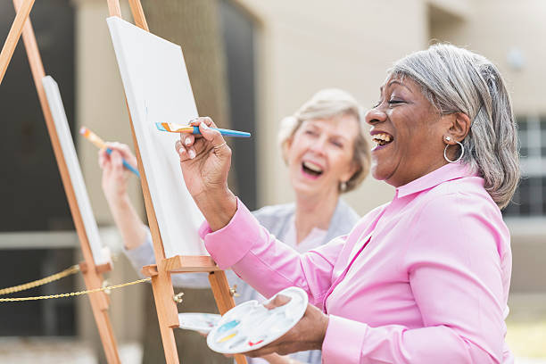 Two senior women having fun painting in art class Two multi-ethnic senior women sitting outdoors at easels painting pictures on canvases. The focus is on the African American woman who is holding a paintbrush and looking up at her artwork as she laughs. leisure activity stock pictures, royalty-free photos & images