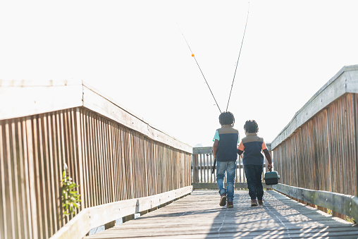 Rear view of two little African American boys going fishing. They are walking on a wooden bridge toward the water carrying rods and a tackle box. It is a bright, sunny day.