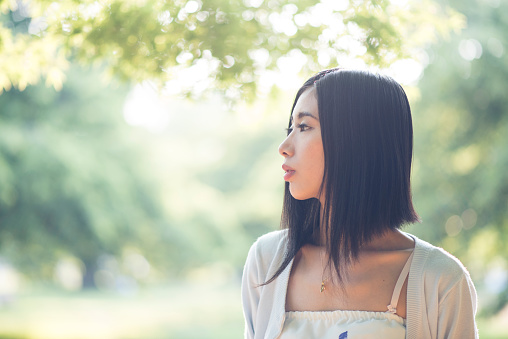 Portrait of a young Japanese woman in Tokyo, Japan. She is smiling and is looking away. Green trees and sun in the back.