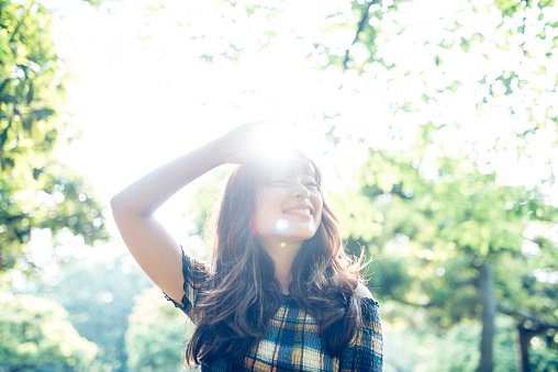 Portrait of a young Japanese woman in Tokyo, Japan. She is smiling, holding her hand on her head and is looking at camera. Green trees and sun in the back.