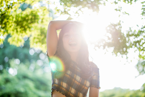Portrait of a young Japanese woman in Tokyo, Japan. She is smiling, holding her hand on her head and is looking at camera. Green trees and sun in the back.