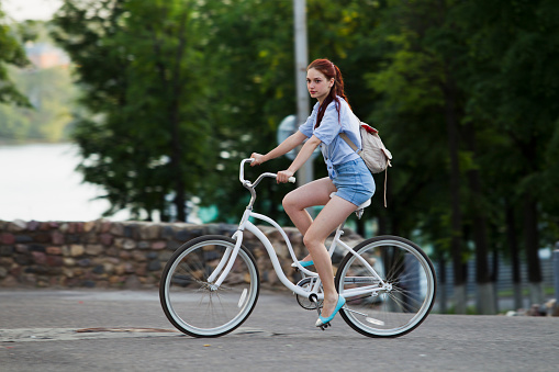 Girl in blue shorts and a denim shirt, riding on asphalt on a white bicycle summer evening in the city, on the background of green trees, stone walls.