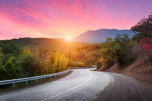 Mountain road passing through the forest with colorful sky and red clouds at sunset in summer. Mountain landscape with road. travel background