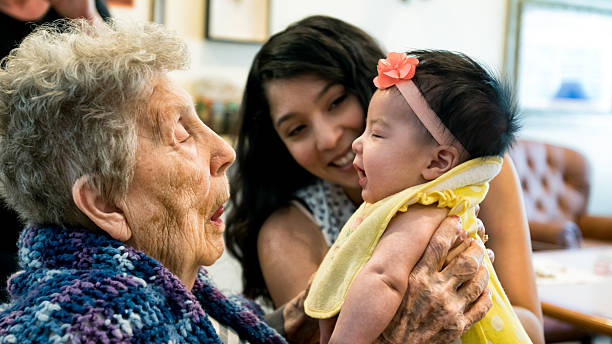 Elderly Woman Holding Infant Granddaughter as Mother Looks On Smiling Beautiful, poignant image of a very old great grandmother holding her beautiful mixed race infant great granddaughter for the very first time as the baby's mother looks on with a big warm smile.  All three people have authentic facial expressions. hispanic grandmother stock pictures, royalty-free photos & images