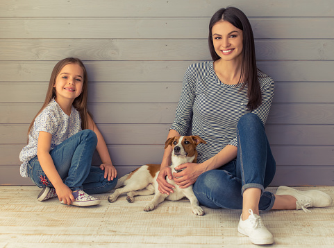 Cute little daughter and her beautiful young mother are looking at camera and smiling while posing with their dog