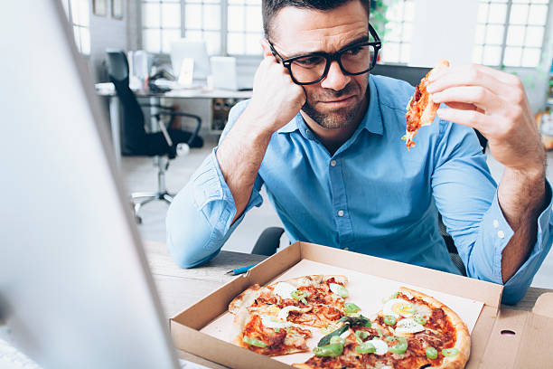 Young businessman having unpleasent pizza lunch break at workplace Young man with eyeglasses and shirt eating pizza at workplace. Man sitting hand on cheek, holding a slice of pizza in hand and looking at it. Pizza box on table. Desk with computers and windows on background. disgust stock pictures, royalty-free photos & images