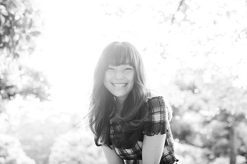 Black and white portrait of a young Japanese woman in Tokyo, Japan. She is smiling, holding her hand behind her head and is looking at camera. Trees in the back.